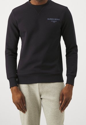 Mand iført en sort Björn Borg sweatshirt med blå logotekst på brystet og lysegrå joggingbukser, stående med hænderne afslappet ned langs siderne.