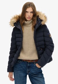 Woman with long hair wearing a beige knitted turtleneck, navy blue puffer jacket with fur hood, blue jeans, and braided brown belt, hand in pocket.