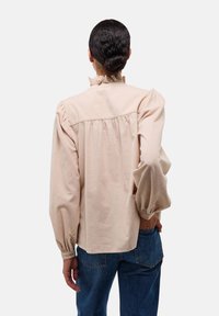 Blouse beige clair avec un col volanté et des manches ballons, en tissu texturé, avec un détail froncé dans le dos, portée avec un jean bleu.