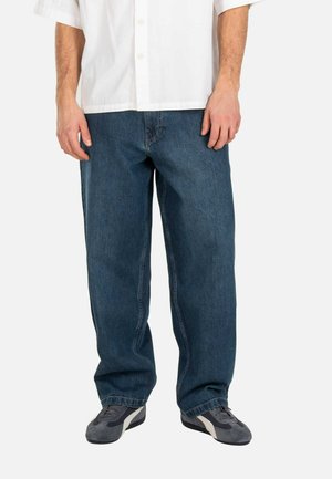 Mann trägt lockere blaue Jeans, graue Turnschuhe und ein weißes kurzärmeliges Hemd mit Knöpfen, steht vor einem schlichten Hintergrund.