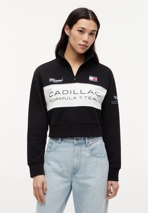 Nő fekete, félcipzáras pulóverben, amelyen a „Cadillac Formula 1 Team” felirat és a Miami logó látható, világoskék, magasderekú farmernadrágban.