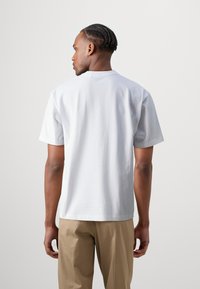 Weißes Baumwoll-T-Shirt mit kurzen Ärmeln, Rundhalsausschnitt und geradem Schnitt, getragen mit beigen Hosen. Einfaches Design, keine sichtbaren Muster.