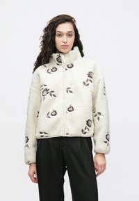 Jakna od flisa u boji kreme s visokim ovratnikom, ukrašena smeđim cvjetnim vezom. Prednji dio s punim patentnim zatvaračem i opuštenim krojem. Glatka tekstura.