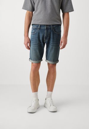 Homme portant un short en jean bleu retroussé, un t-shirt gris, des chaussettes blanches et des baskets blanches, debout devant un fond uni.