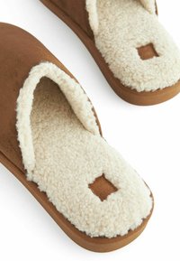 Rudos suede šlepetės su minkšta grietinėlės spalvos avikailio pamušalu ir tekstūruota gumine padanga, su kvadratiniu logotipo ženkleliu ant pirštų.