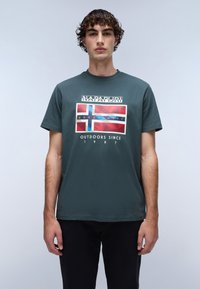 Zelené bavlněné tričko s barevným grafickým designem s motivem vlajky, textem "NAPAPURRI" a "OUTDOORS SINCE 1987" uprostřed.