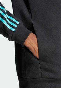 Svart sweatshirt med turkosa tre-stripesdesign på ärmen. Har en sidoficka och mjuk tygkänsla.