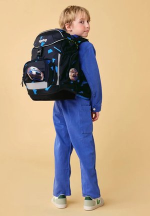Boy in blue outfit wearing black and blue backpack with car and skateboard patches, standing against beige background.