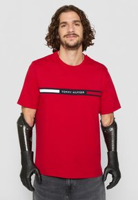 Küljelt lokkis juustega mees, kes kannab punast Tommy Hilfiger T-särki ja musti süsinikkiust proteesi eelkäsi koos kinnastega, seisab lihtsa tausta ees.