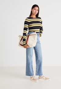 Canvas tote bag med bruna handtag, krämfärgad, stor storlek. Modell som bär en grön och svart randig ribbad topp, ljusblå raka jeans, beige sneakers.