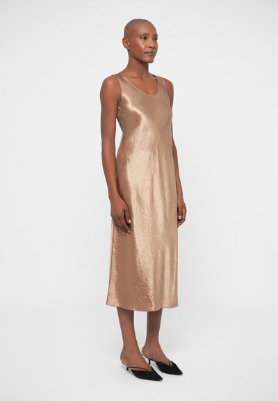 Μπρονζέ σατέν φόρεμα με στρογγυλή λαιμόκοψη, χωρίς μανίκια και μήκος μέχρι το γόνατο. Συνδυάζεται με μαύρα παπούτσια με μυτερή μύτη. Λείο υλικό.