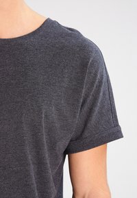 Camiseta de algodón gris con mangas cortas enrolladas, un cuello redondo y una textura suave. Detalle de costura visible en el dobladillo.