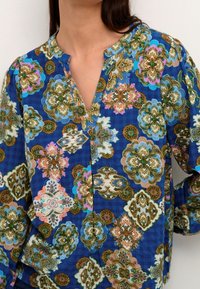 Kobieta nosząca niebieską bluzkę w wzory, z kwiatowymi i geometrycznymi motywami, z dekoltem w kształcie litery V i długimi rękawami.