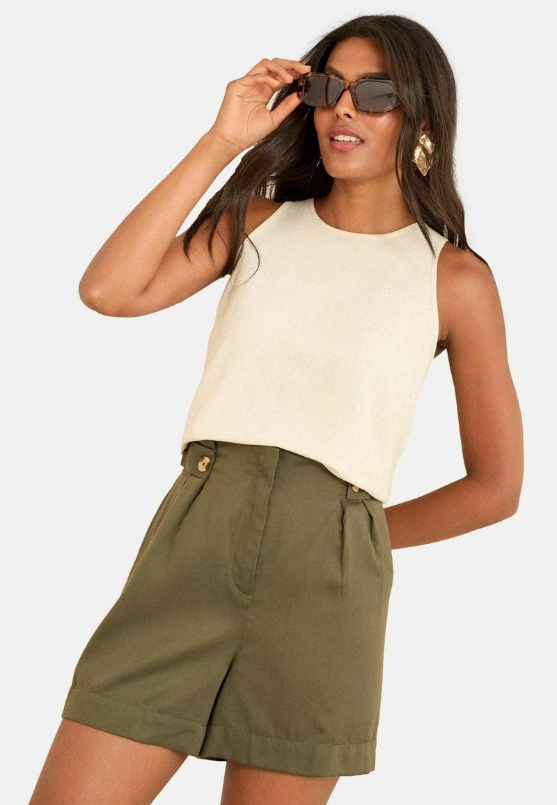 Ljuscremefärgad ärmlös topp i kombination med olivgröna höga shorts med knappar, visad med solglasögon och långt hår.