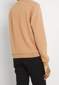 Una persona con el pelo corto lleva un suéter suave de color marrón claro, de espaldas, mostrando un tatuaje en su mano y unos pantalones vaqueros negros.