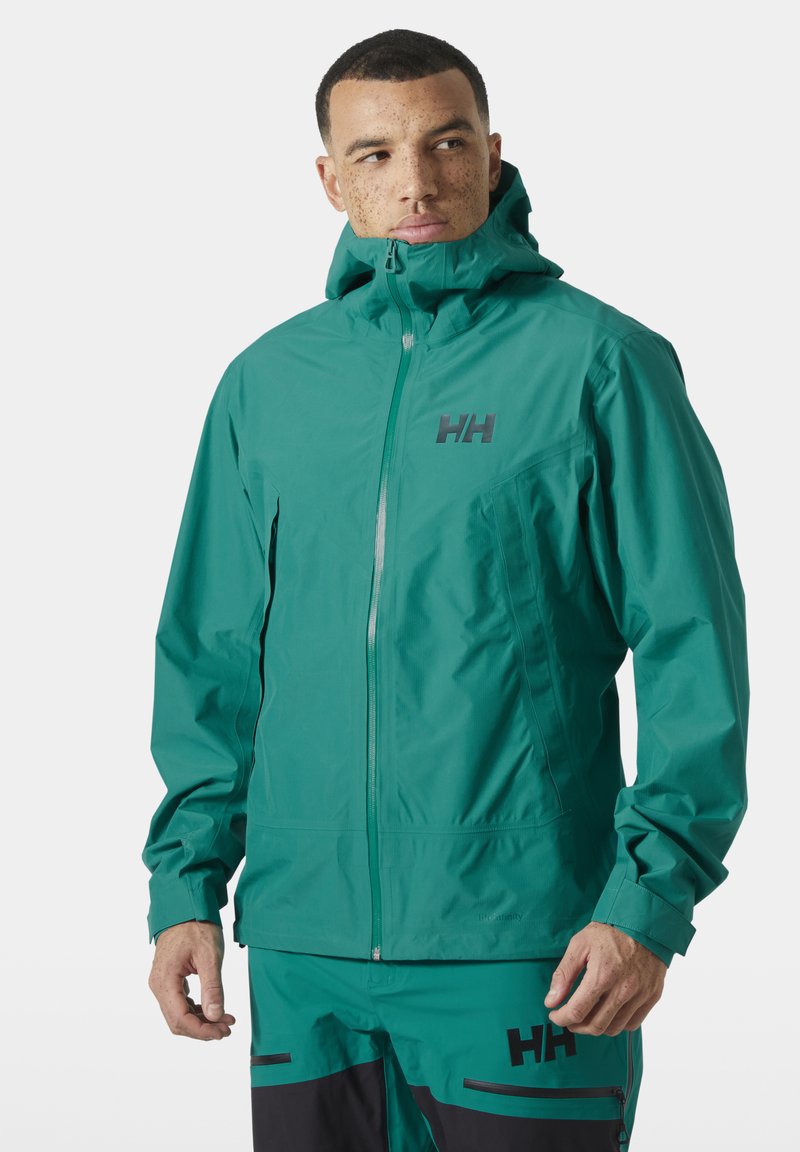 Man draagt een waterdichte jas met volledige rits en capuchon in teal en bijpassende broek, beide met het "HH"-logo, kijkt naar de zijkant.