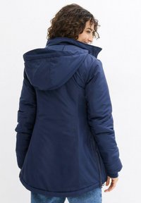 Cappotto imbottito con cappuccio di colore blu navy, con vita arricciata, dettagli con zip e una texture liscia. Presenta un orlo arrotondato e polsini elasticizzati.