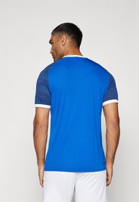 Maglietta sportiva blu a maniche corte, con parte posteriore uniforme e dettagli a fantasia blu sulle spalle. Bordo bianco sul collo.