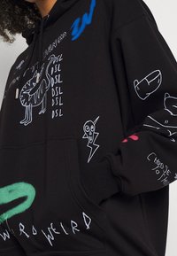 Sudadera negra con garabatos en blanco, que incluyen un perro, una calavera con un rayo, una mano y texto en estilo graffiti como "DSL" y "WEIRD."