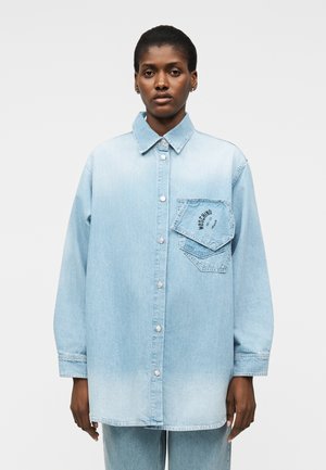 Mustanahaline inimene, kes kannab liialdatult suurt helesinist teksariidest särk, kus on suur tasku mustriga "Moschino Jeans", ning sobivaid teksapükse.