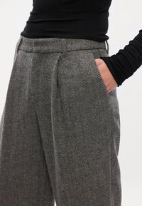 Calças de padrão espinha de peixe cinza, com cintura alta, pregas e bolsos laterais. O tecido é texturizado, proporcionando uma aparência estruturada.