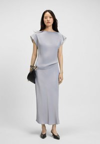 Hellgraues Maxikleid mit kurzen Ärmeln, glatter Textur und figurbetonter Silhouette; kombiniert mit einer schwarzen Handtasche und schwarzen High-Heel-Mules.