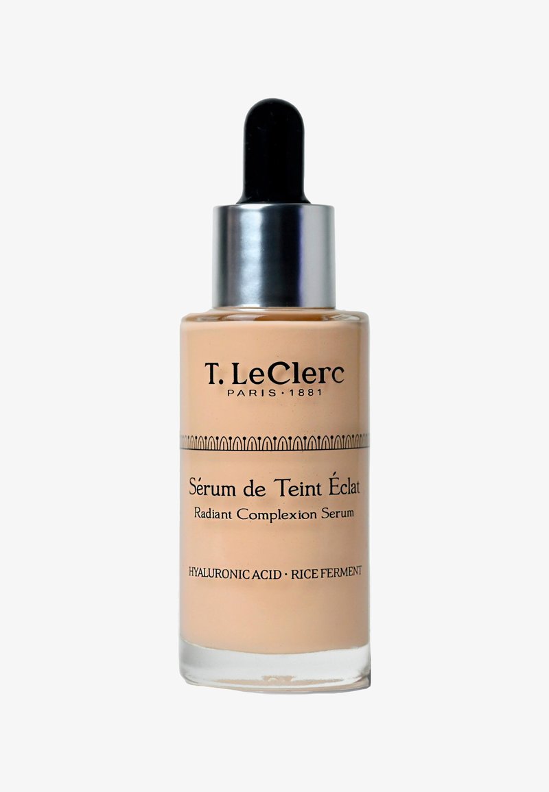 Bottiglia contagocce in vetro di siero per un incarnato radioso, liquido beige chiaro, tappo argentato, testo nero e fascia decorativa a motivo.