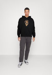 Hanorac negru cu glugă, cu un design grafic circular pe piept, combinat cu pantaloni cargo gri și adidași negri cu glezna înaltă.