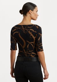 Svart topp med brunt kjede-design, med vid hals og trekvartlange ermer. Modellen har på seg svarte bukser og et svart belte.