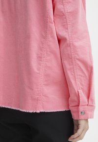 Różowa dżinsowa koszula z długimi rękawami, postrzępionym dołem i detalem na mankietach z napami. Charakteryzuje się delikatną fakturą i kontrastowym przeszyciem.