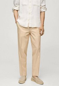 Pantalons beige ajustés en tissu léger, avec une coupe droite et des poches sur les côtés, associés à une chemise blanche à rayures avec boutonnières.