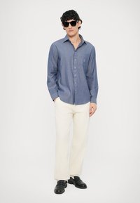 Jasnoniebieska koszula na guziki z długim rękawem, zestawiona z kremowymi spodniami o szerokich nogawkach i czarnymi butami, noszona z okularami przeciwsłonecznymi.