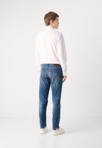 White long-sleeve shirt, blue jeans, light texture, slim fit, visible stitching, brown leather label on the waistband, white sneakers.