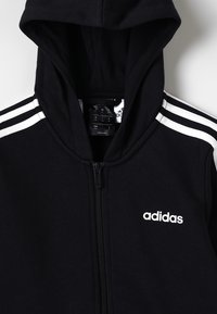 Felpa nera Adidas con zip, caratterizzata da una chiusura frontale, logo bianco sul petto e dettagli a strisce bianche sulle maniche. Materiale morbido.