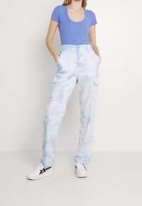 Woman wearing a blue ribbed short-sleeve top, light blue tie-dye cargo pants, and white sneakers with black stripes, standing with hands in pockets.