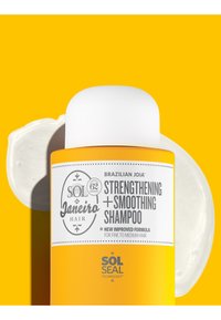 Botella amarilla del champú brasileño Joia fortalecedor y suavizante con tapa blanca sobre un fondo amarillo con una mancha de crema blanca detrás.