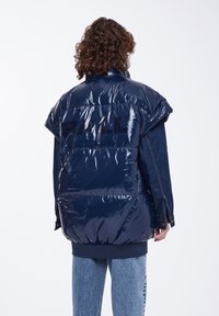 Abrigo acolchado de color azul marino con acabado brillante, diseño oversized y hombros marcados. Presenta una manga de denim oscuro y un logo en la parte posterior.