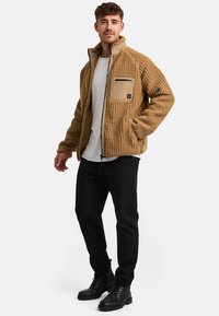 Chaqueta de forro polar texturizado marrón con un alto cuello, cierre de cremallera, y un bolsillo en el pecho en color beige en contraste, combinada con pantalones negros de corte slim y botas.