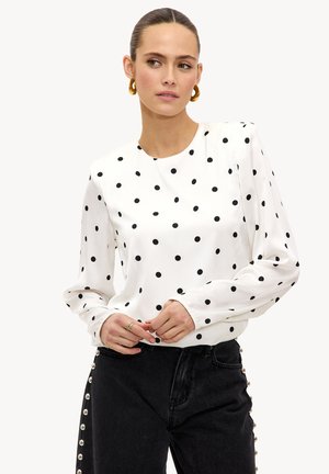 Blouse blanche à pois noirs, manches longues et encolure ronde. Associée à un jean noir taille haute avec des accents en perles.