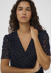 Tengerészkék ruha rövid, áttetsző ujjal, texturált virágmintával és mély V-nyakkivágással. Simább anyag finom fényű felülettel.