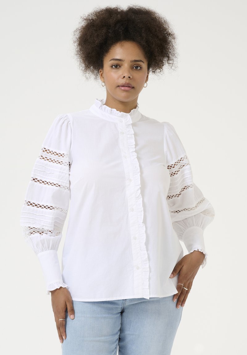 Chemise blanche avec col volanté et devant froncé, dotée de manches bouffantes avec inserts en dentelle, réalisée en tissu lisse, accompagnée d'une coupe décontractée.