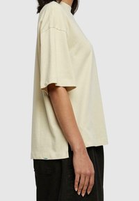 Camiseta de color crema, de manga corta, hecha de algodón suave. Corte relajado con cuello redondo y una pequeña etiqueta verde en el dobladillo.