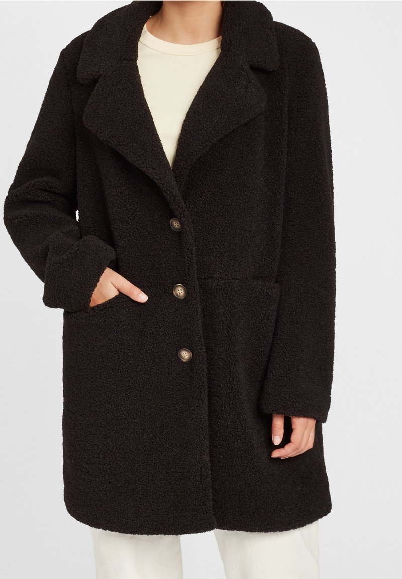 Femme portant un manteau noir texturé mi-long avec trois boutons et un large col, main dans une poche, sur un haut blanc et un pantalon.