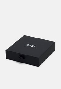 Schwarze rechteckige Box mit einer glatten Textur und einer herausziehbaren Schublade. Auf der Oberseite befindet sich das Wort "BOSS" in Weiß, zentriert.