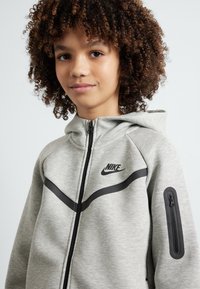 Šedý mikina na zips Nike s kapucňou, čiernym logom a diagonálnymi čiernymi akcentmi. Má bočnú vrecko so zipsom a textúrovanú tkaninu.
