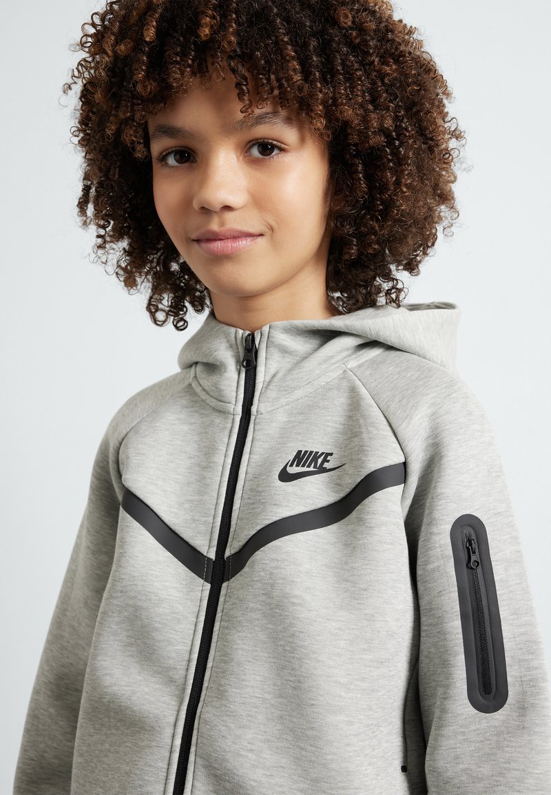 Šedý mikina na zips Nike s kapucňou, čiernym logom a diagonálnymi čiernymi akcentmi. Má bočnú vrecko so zipsom a textúrovanú tkaninu.