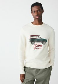 Noor meester, kes kannab valget sviitrit, millel on Ford Bronco auto graafika, ja oliivrohelisi pükse, seistes lihtsa valge taustaga.