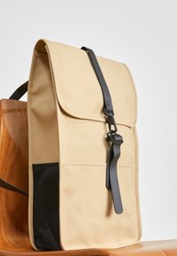 Beige ryggsäck med en strukturerad form, matt finish och svart rem. Har ett klaffstängning och en sidoficka med en texturerad tyg.