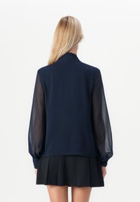 Morska bluza z prosojnimi dolgimi rokavi in visokim ovratnikom. Gladek glavni material kontrastira z teksturiranimi detajli na rokavih.
