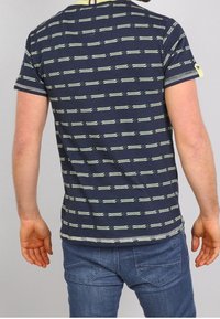 Mies, joka on pukeutunut tummansiniseen raidoitettuun lyhythihaiseen paitaan ja sinisiin farkkuihin, seisoo selin kameraan yksinkertaista vaaleaa taustaa vasten.